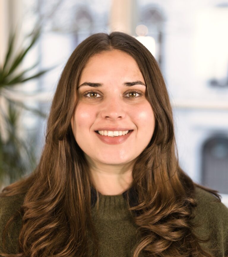 Eine Frau mit langen braunen Haaren lächelt in die Kamera. Sie trägt einen dunkelgrünen Pulli