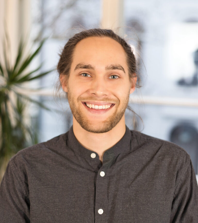 Ein Mann mit blonden Haaren lächelt in die Kamera. Er trägt ein graues Hemd.