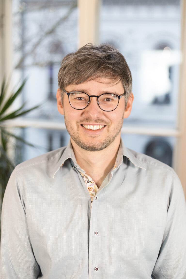 Ein Mann mit Brille lächelt in die Kamera. Er trägt ein helles Hemd.