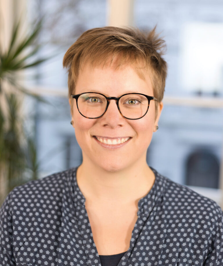 Eine Frau mit kurzen blonden Haaren und dunkler Brille lächeltin die Kamera. Sie trägt eine blaue Bluse.