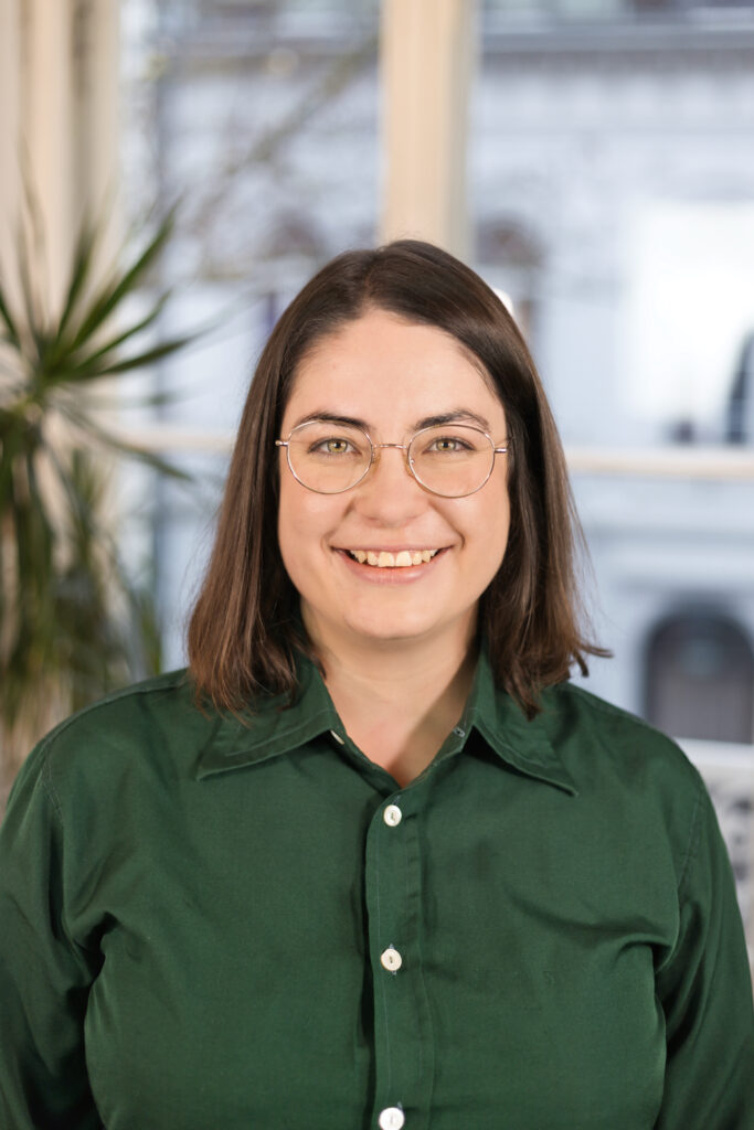 Eine Frau mit schwarzen Haaren und einer Brille lächelt in die Kamera. Sie trägt eine grüne Bluse.