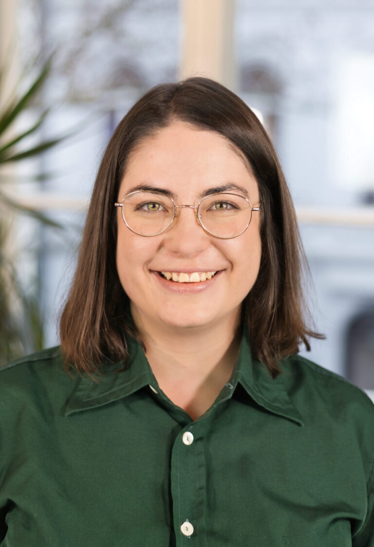 Eine Frau mit schwarzen Haaren und einer Brille lächelt in die Kamera. Sie trägt eine grüne Bluse.