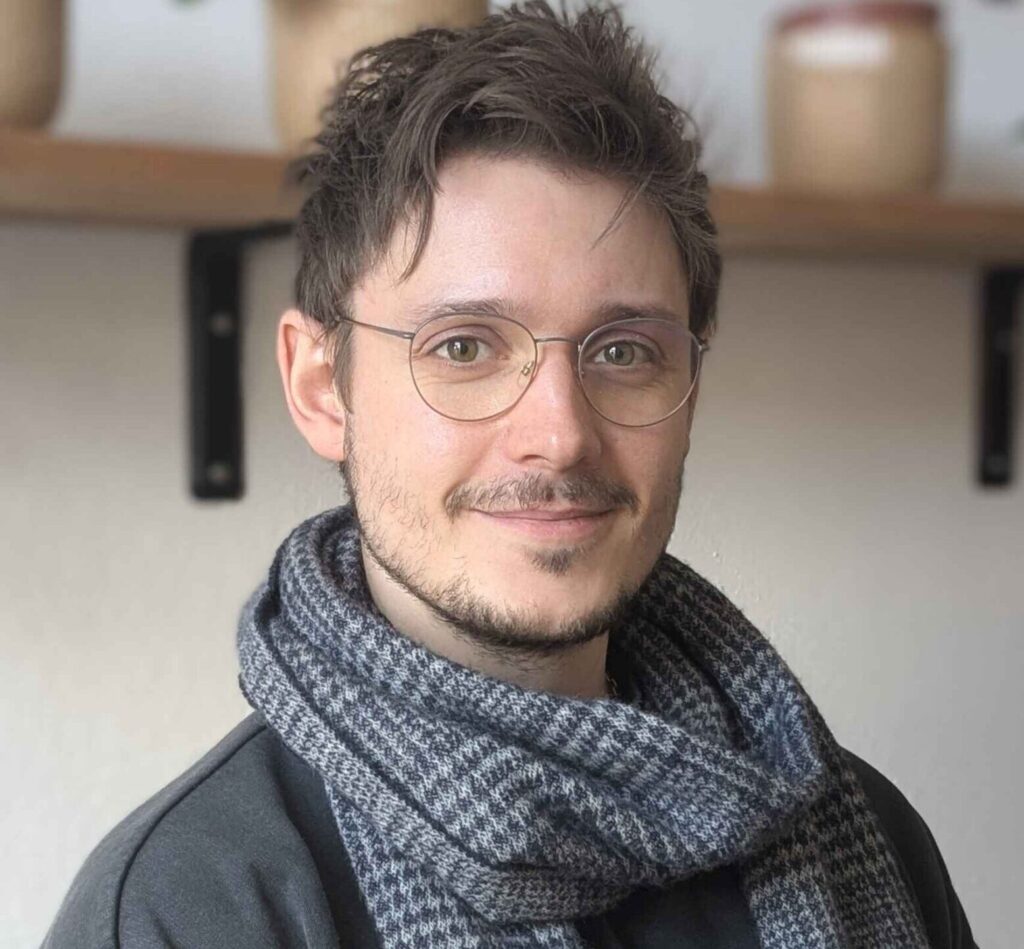 Ein Mann mit Brille und Bart und dunklen Haaren lächelt in die Kamera. Er trägt einen grauen Pulli und einen grauen Schal.