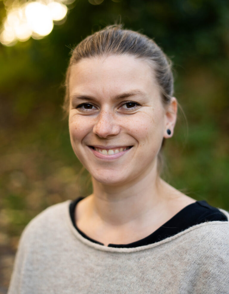 Eine Frau mit blonden Haaren lächelt in die Kamera. Sie trägt einen grauen Pulli.