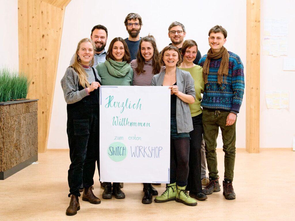 Neun Menschen stehen in einer Gruppe und lächeln in die Kamera. Zwei halten gemeinsam ein Plakat hoch mit der Aufschrift "Herzlich Willkommen zum SWITCH-Workshop"
