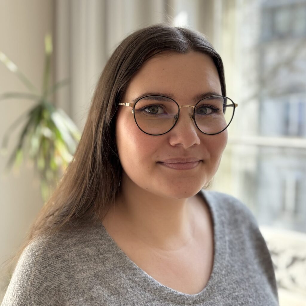 Eine Frau mit brauen Haaren lächelt in die Kamera. Sie trägt eine Brille und einen grauen Pulli.