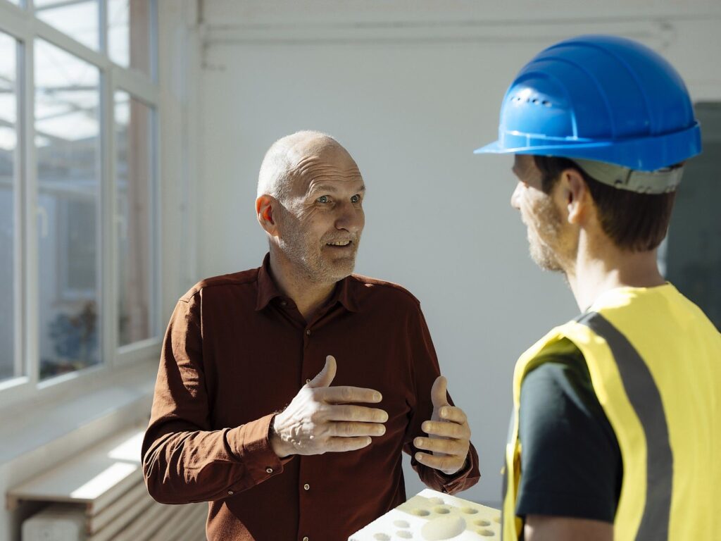 Ein Mann im roten Hemd erklärt einem Mann mit blauem Helm etwas.