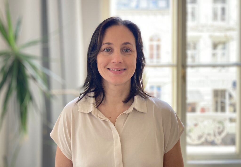 Eine Frau mit dunklen Haaren und heller Bluse lächelt in die Kamera.