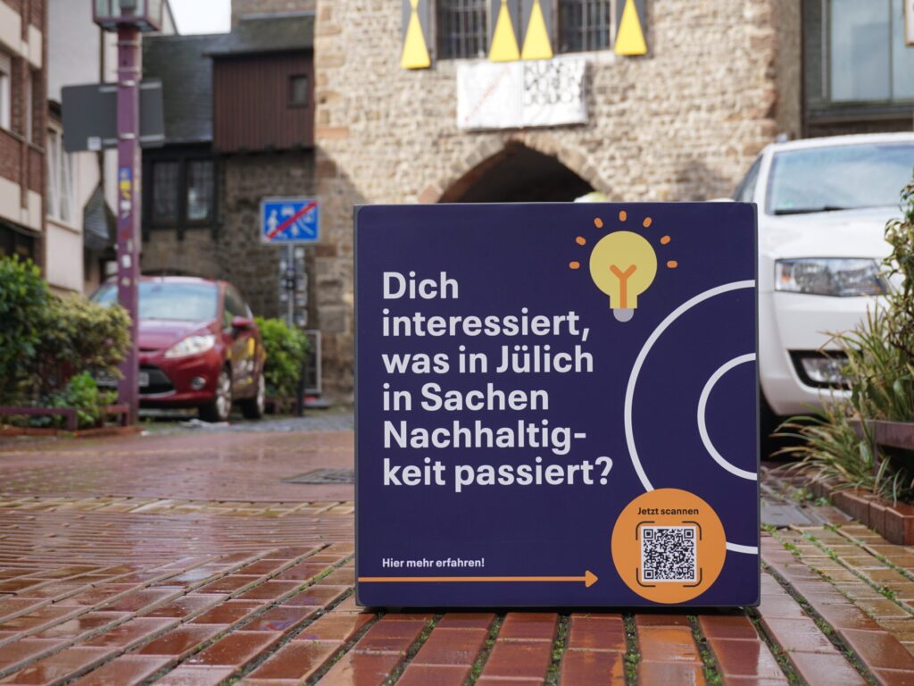Ein blauer, großer Würfel steht auf einem Parkplatz. Auf dem Würfel steht "Dich interessiert, was in Jülich in Sachen Nachhaltigkeit passiert?"