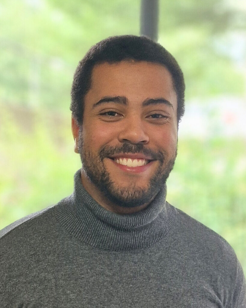 Man sieht einen jungen Mann, der in die Kamera lächelt. Er hat dunkle kurze Haare und er trägt einen grauen Rollkragenpullover. Im Hintergrund ist unscharf ein grüner Baum zu sehen.