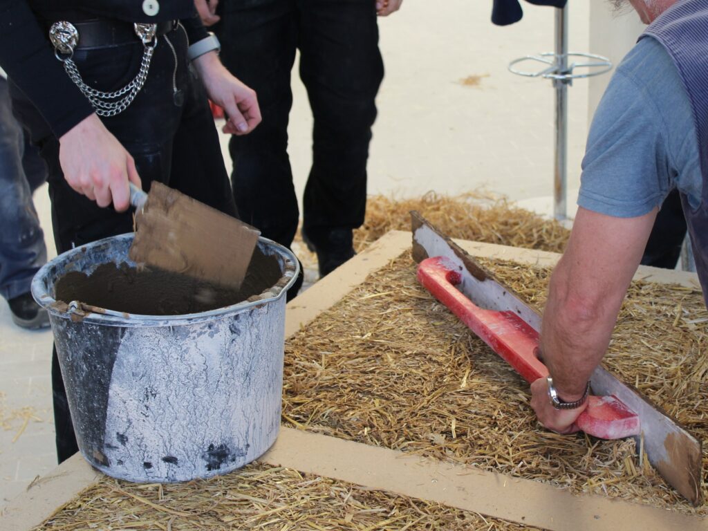 Das Bild zeigt Handwerker*innen bei der Arbeit. Links rührt eine Person einen Eimer Lehmputz an.