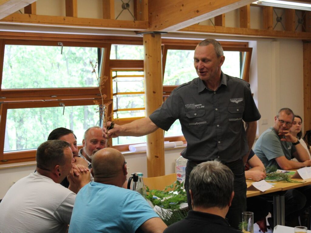 Ein Mann hält einen Ast in der Hand und zeigt ihn Menschen, die an Tischen sitzen.